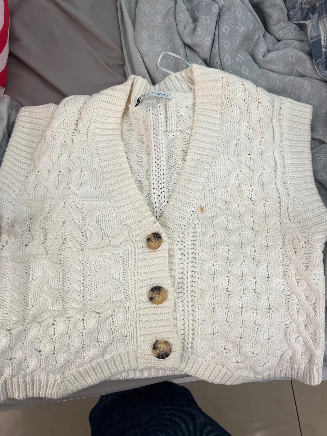 White knitted waistcoat with brown buttons on a bed with grey sheets and a denim garment in the background.