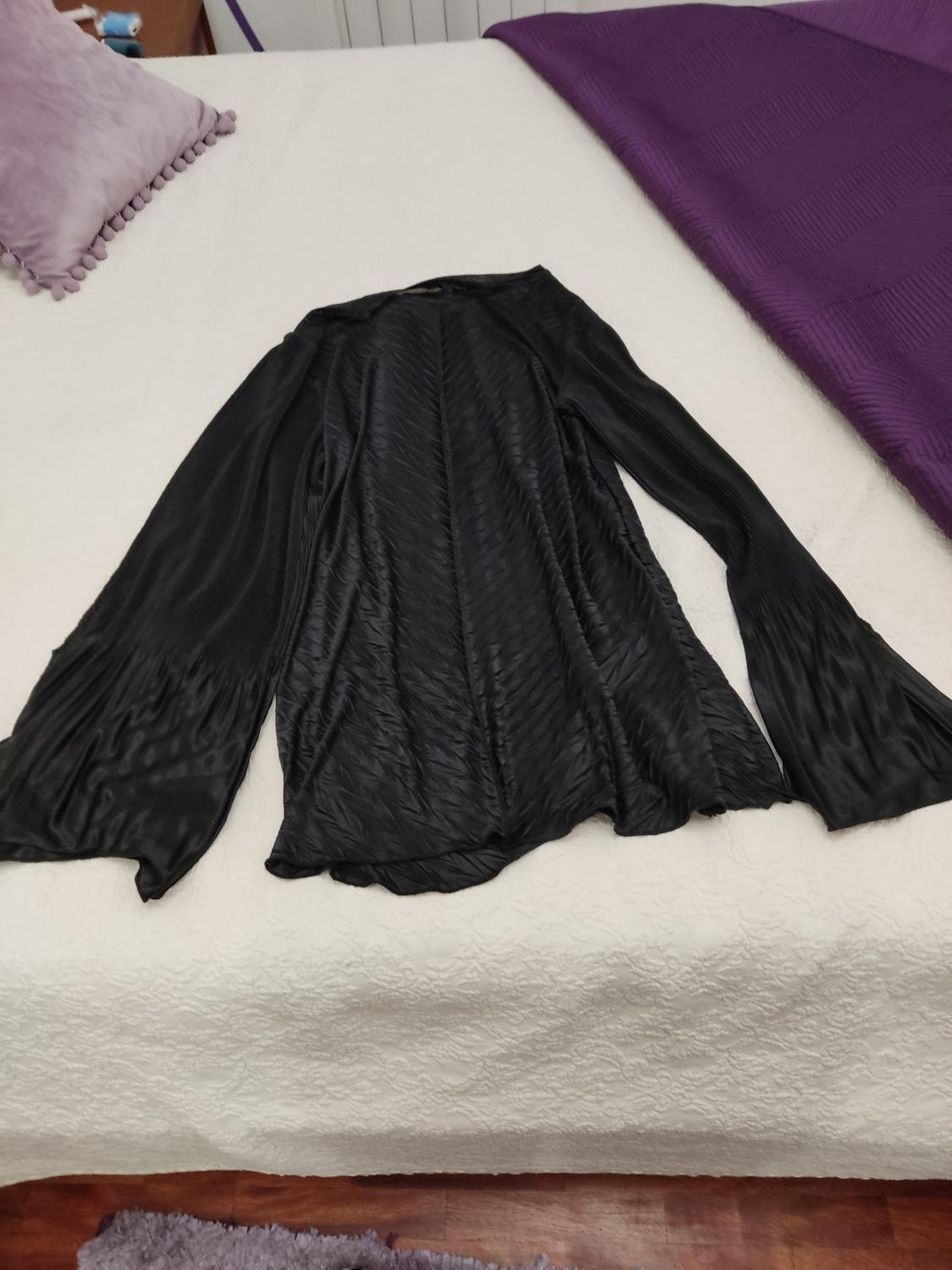 Black long-sleeved blouse with pleated texture on a white bed, with a lilac cushion and a purple blanket in the background.