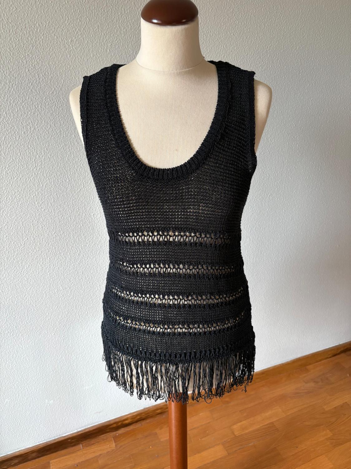 Mannequin wearing a black sleeveless knitted top with a round neck, horizontal openwork stripes and fringing at the hem, on a wooden floor.
