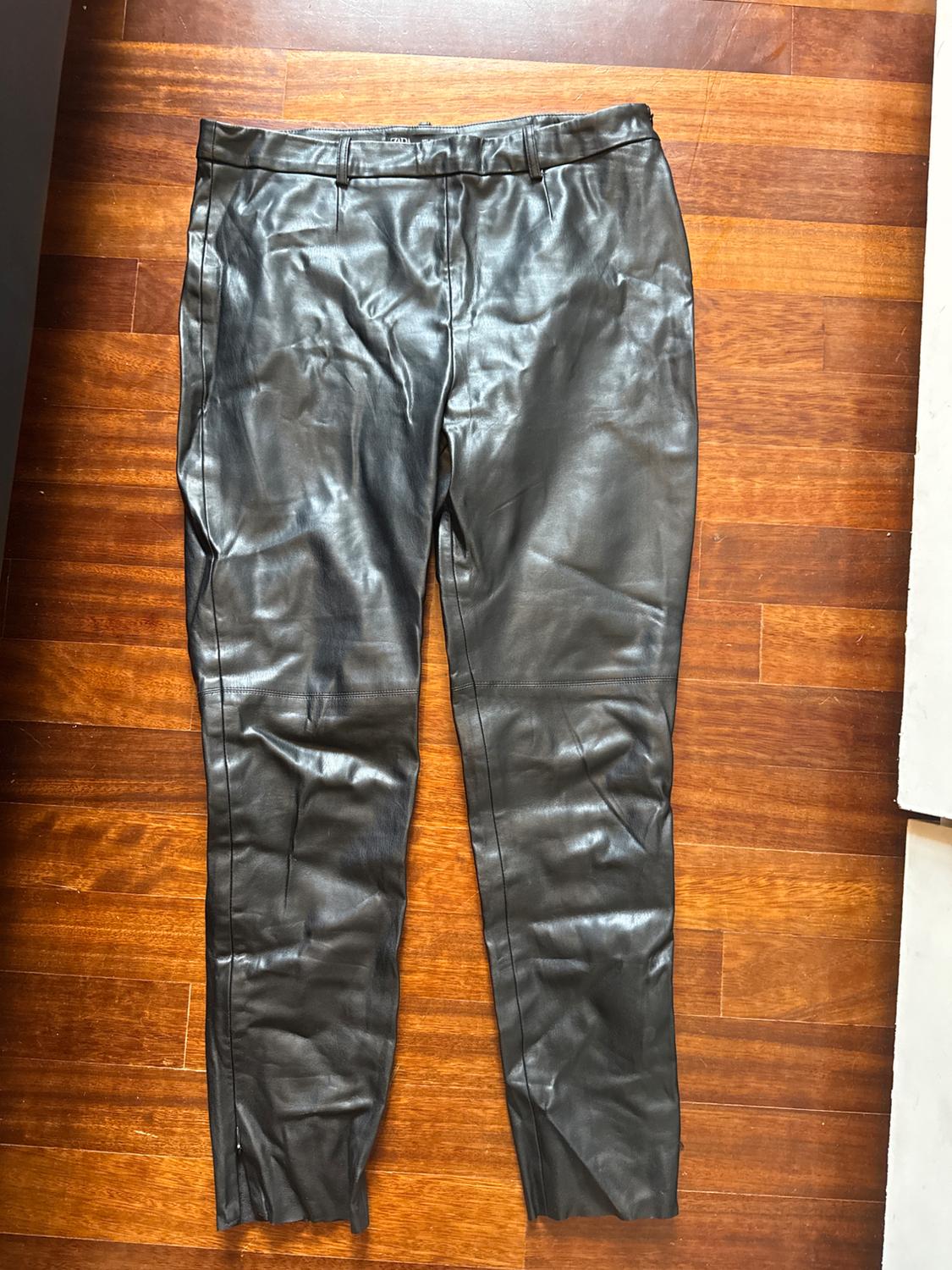 Black leather trousers laid out on a brown wooden floor, displaying a classic design with pockets and belt loops.