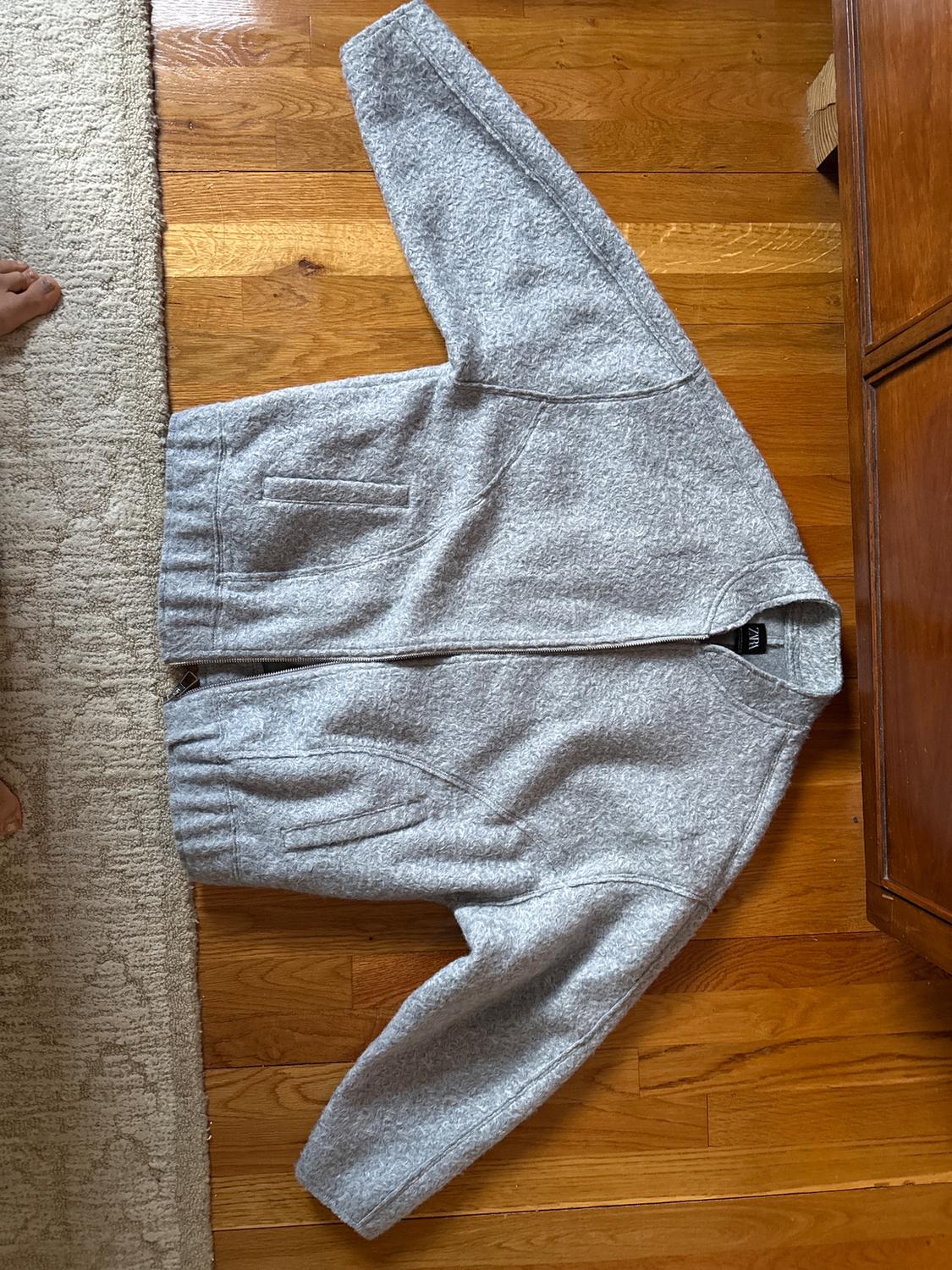 Gray wool zip-up jacket, spread out on a wooden floor next to a light-colored rug.