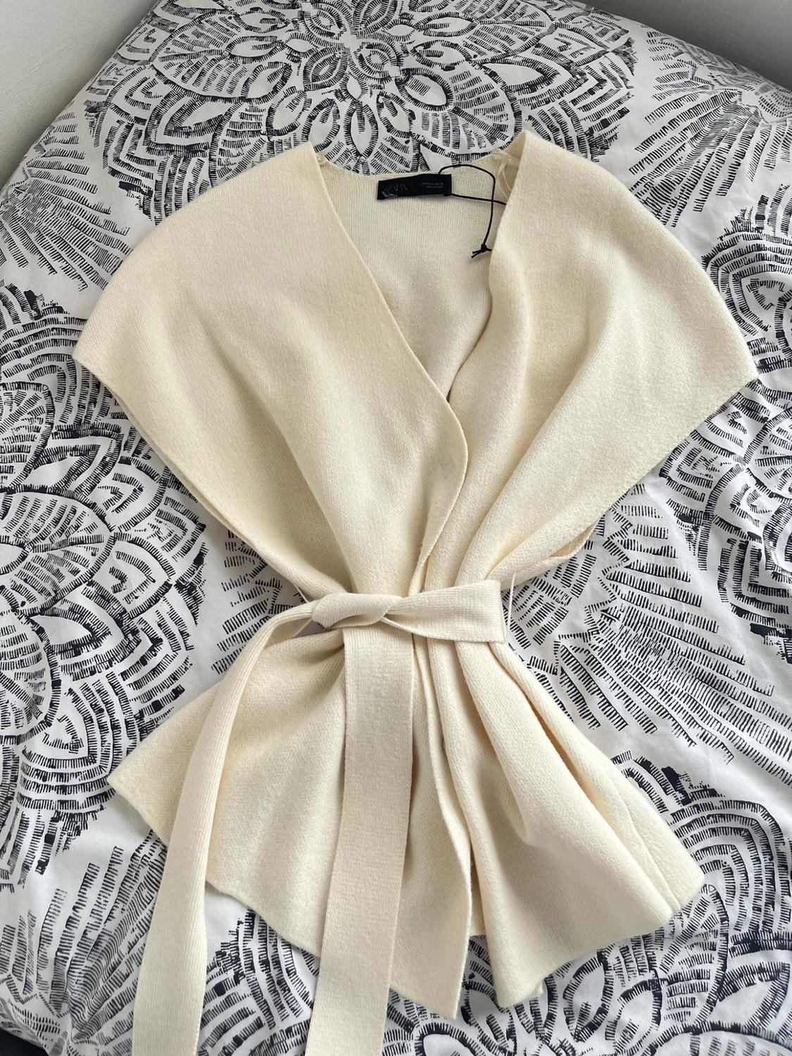 Cream knitted top in a cream colour with a tied belt, against a fabric background with a black-and-white print. Visible text: ZARA.