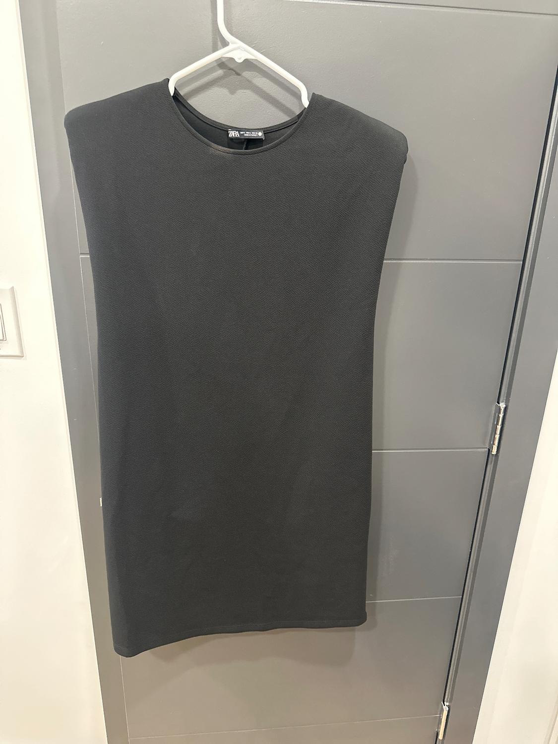 Sleeveless black dress hanging on a white hanger over a gray door. Label visible on the collar.