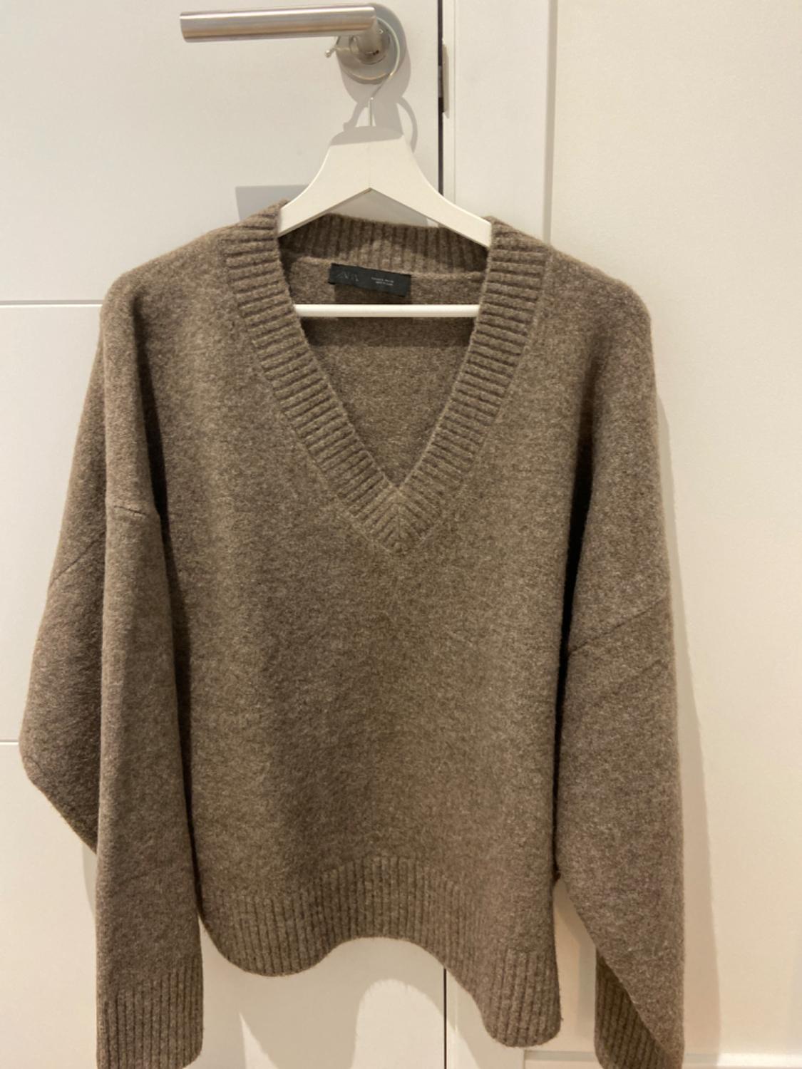 Chunky brown V-neck jumper, hanging on a white hanger over a white door. Label visible on the collar.