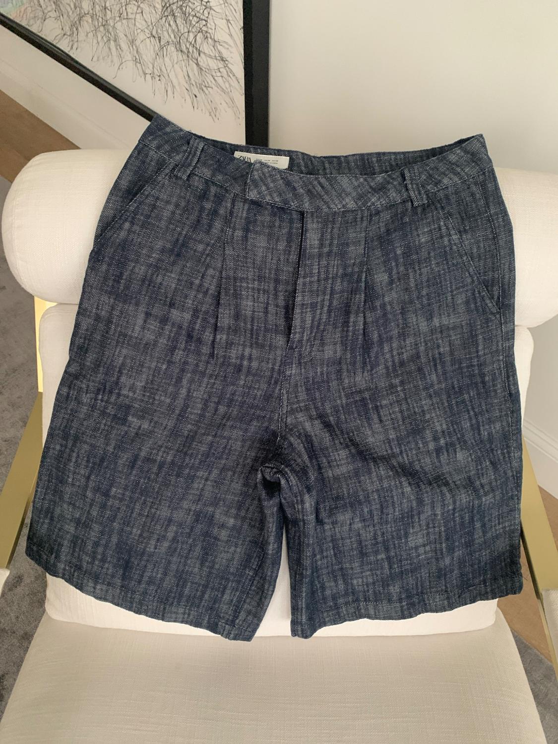 Dark blue textured pleated shorts on a modern white chair. ZARA label visible on the waistband.