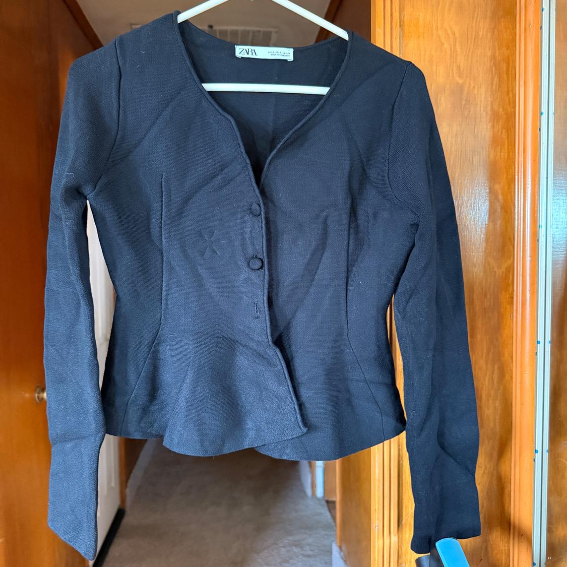 Black long-sleeved jacket hanging on a white hanger, with front buttons, in a light wood setting.