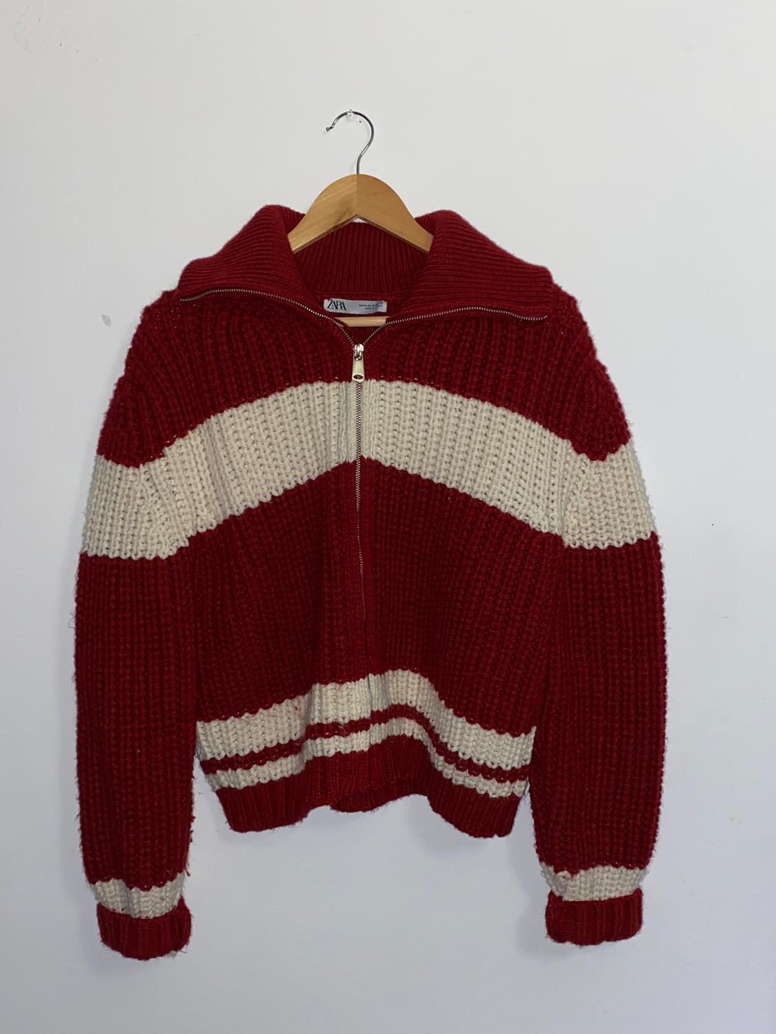 Chunky knit red jumper with white stripes, front fastening and a high neck, hanging on a wooden hanger against a white background.