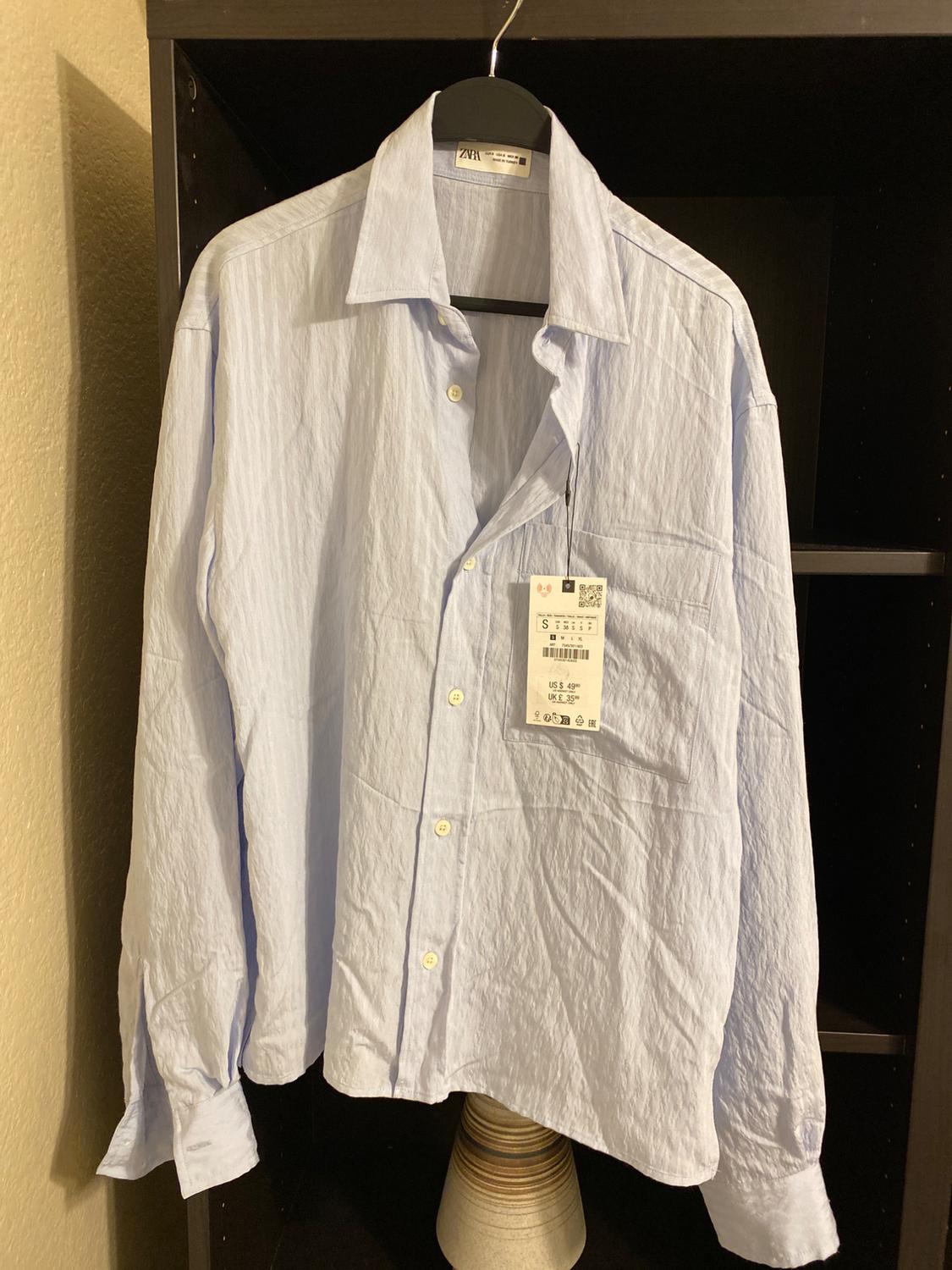 Light blue long-sleeved shirt hanging on a black hanger, with visible tag showing size S and price. Dark shelf background.