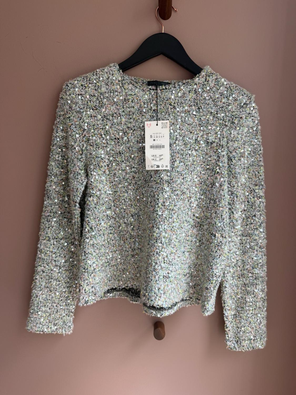Textured sequinned jumper hanging on a black hanger against a pink background. Label with text: S US $49.90 UK £29.99.
