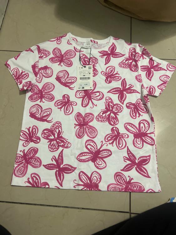 White t-shirt with pink butterfly print, visible hangtag with price of 9.90. Beige tile background.