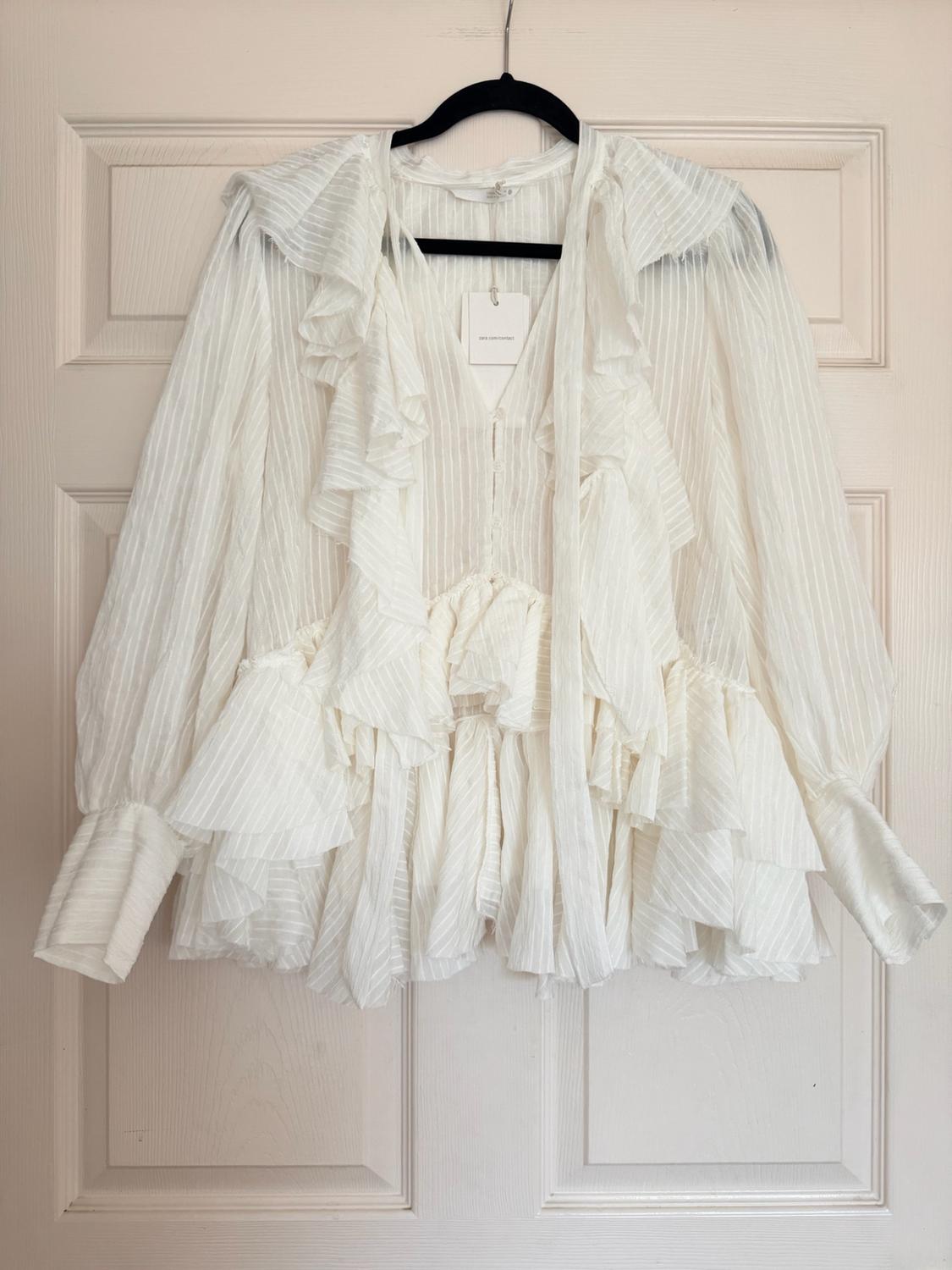 White long-sleeved blouse with ruffles and subtle vertical stripes, hanging on a black hanger on a white door.