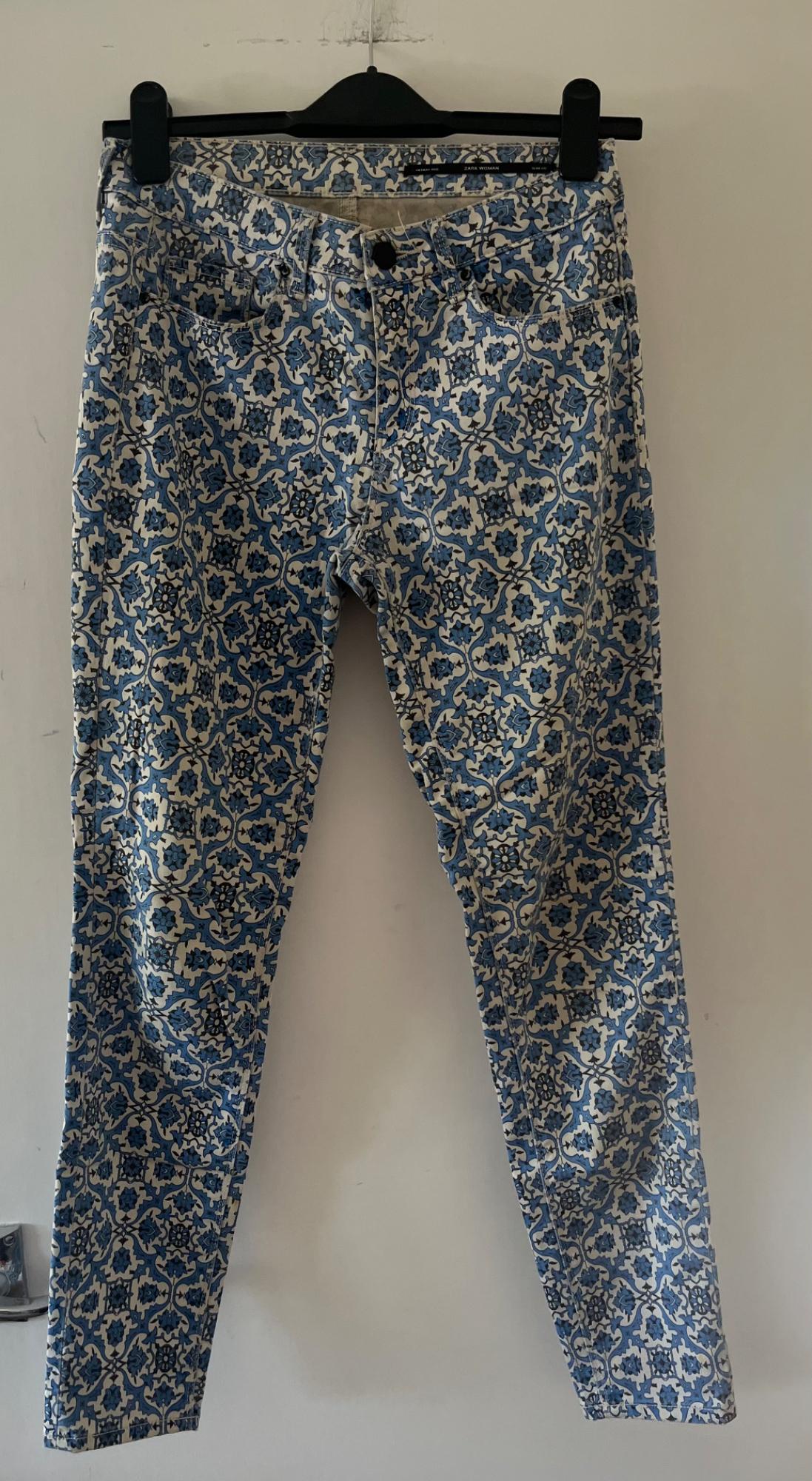 Blue and white floral and geometric print trousers hanging on a hanger against a white wall. Label with the text ZARA WOMAN.