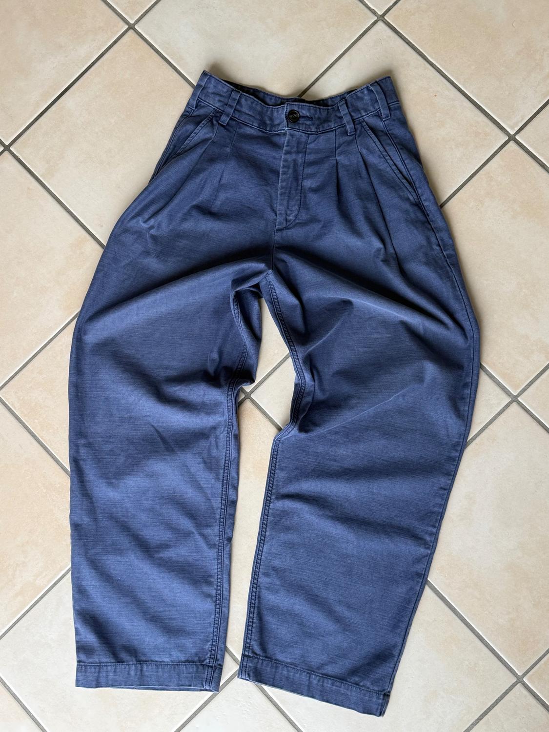 Dark blue, wide-leg trousers with pleats, a black button, and side pockets, on a beige tiled floor.