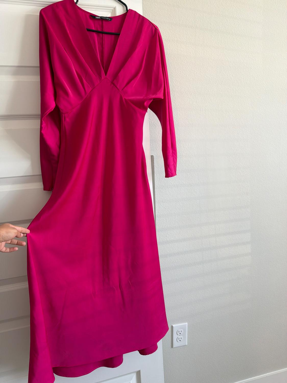 A fuchsia, satin-finish, long-sleeved dress hangs from a white door. A hand is holding the skirt. Inside, there is a label with the text ZARA.