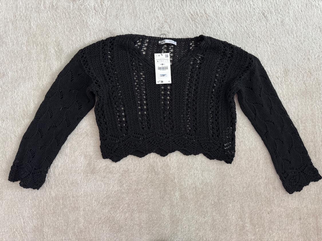 Black crochet jumper with long sleeves and an openwork design, on a light surface. Visible label on the collar.