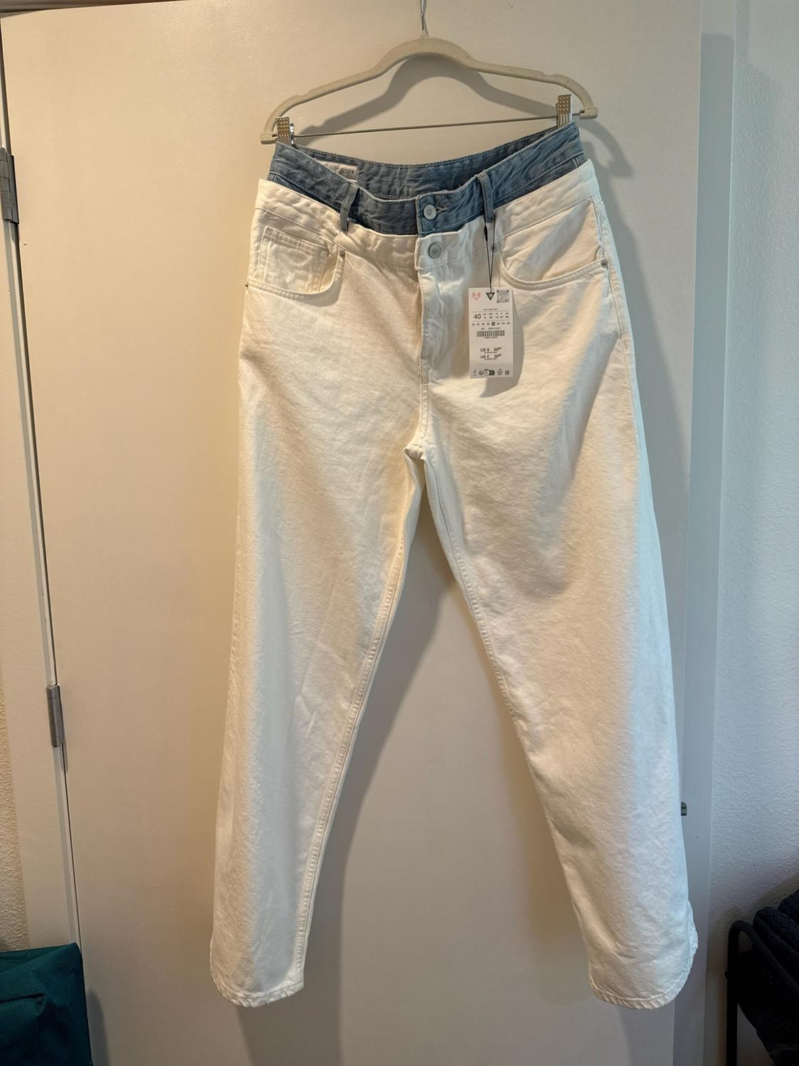 White denim trousers with a blue denim double-waist detail, hanging on a hanger over a white door. It has a label.