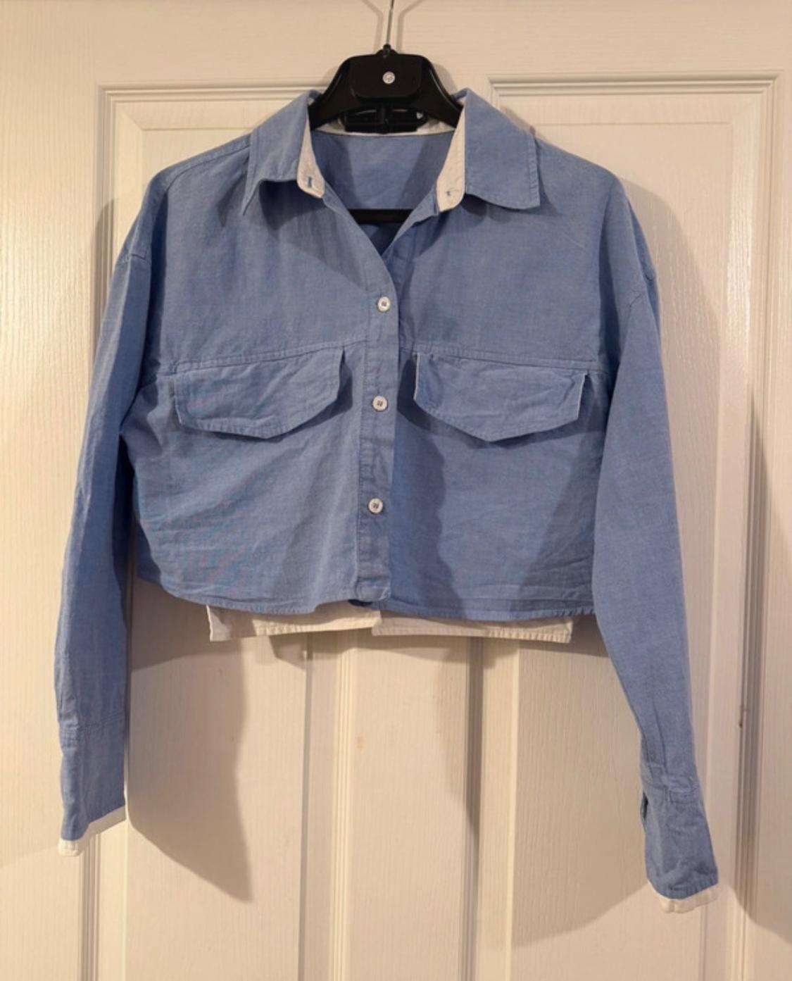 Light blue, long-sleeved, cropped shirt, hanging on a black hanger over a white door.