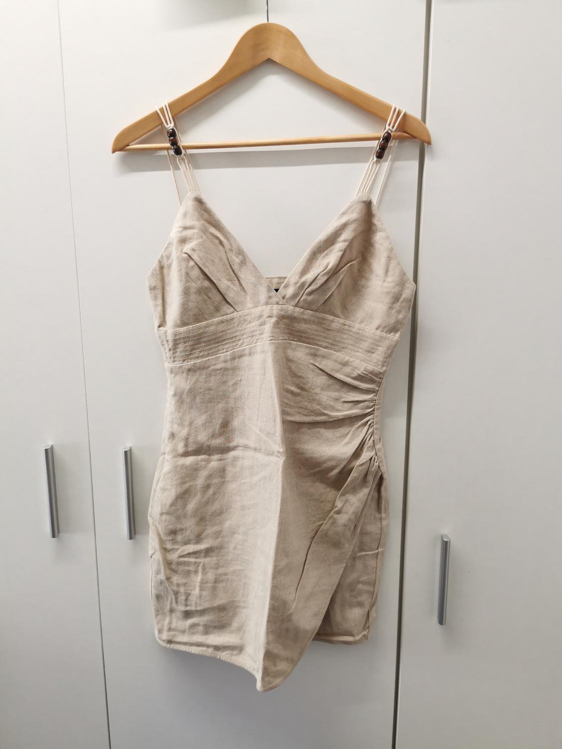 Beige ruched mini dress with a V-neck and wooden detail straps, hanging on a hanger in front of a white wardrobe.