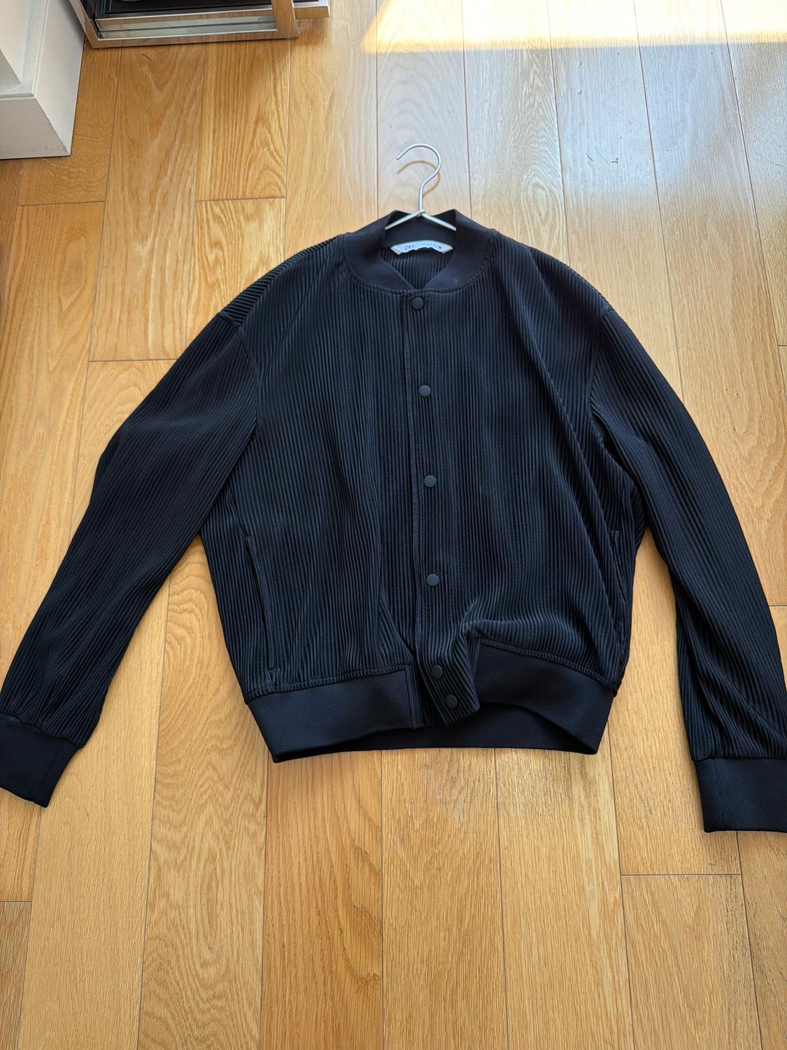Black ribbed fabric jacket with front buttons, hanging on a hanger over a light wood floor.