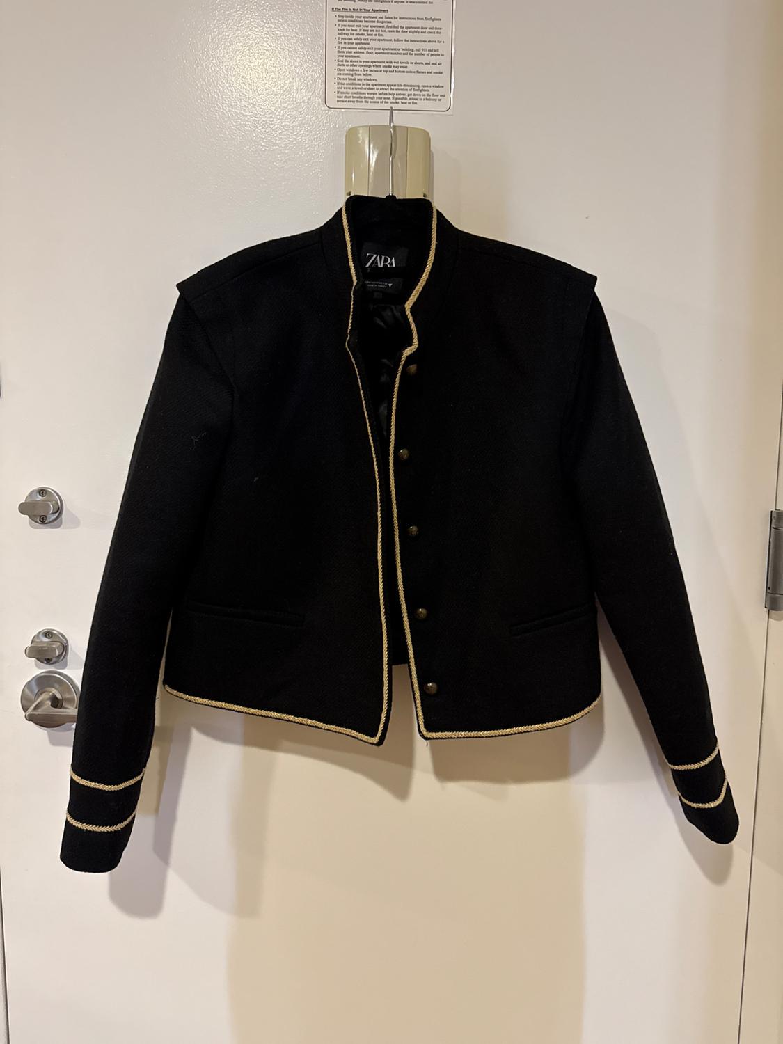 Black jacket with gold trim hanging on a white door. Visible label with the text "ZARA" inside.