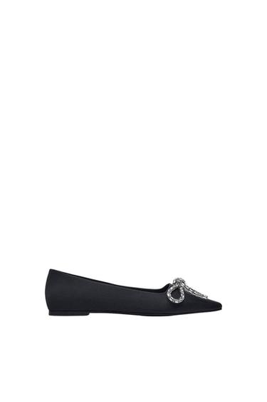 Black ballet flat with silver bow embellishment on the toe, over white background.