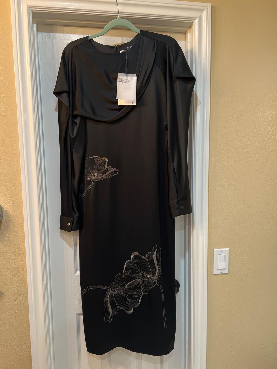 Long-sleeved black dress with a floral design on the bottom, hanging on a white door. Visible label on the collar.