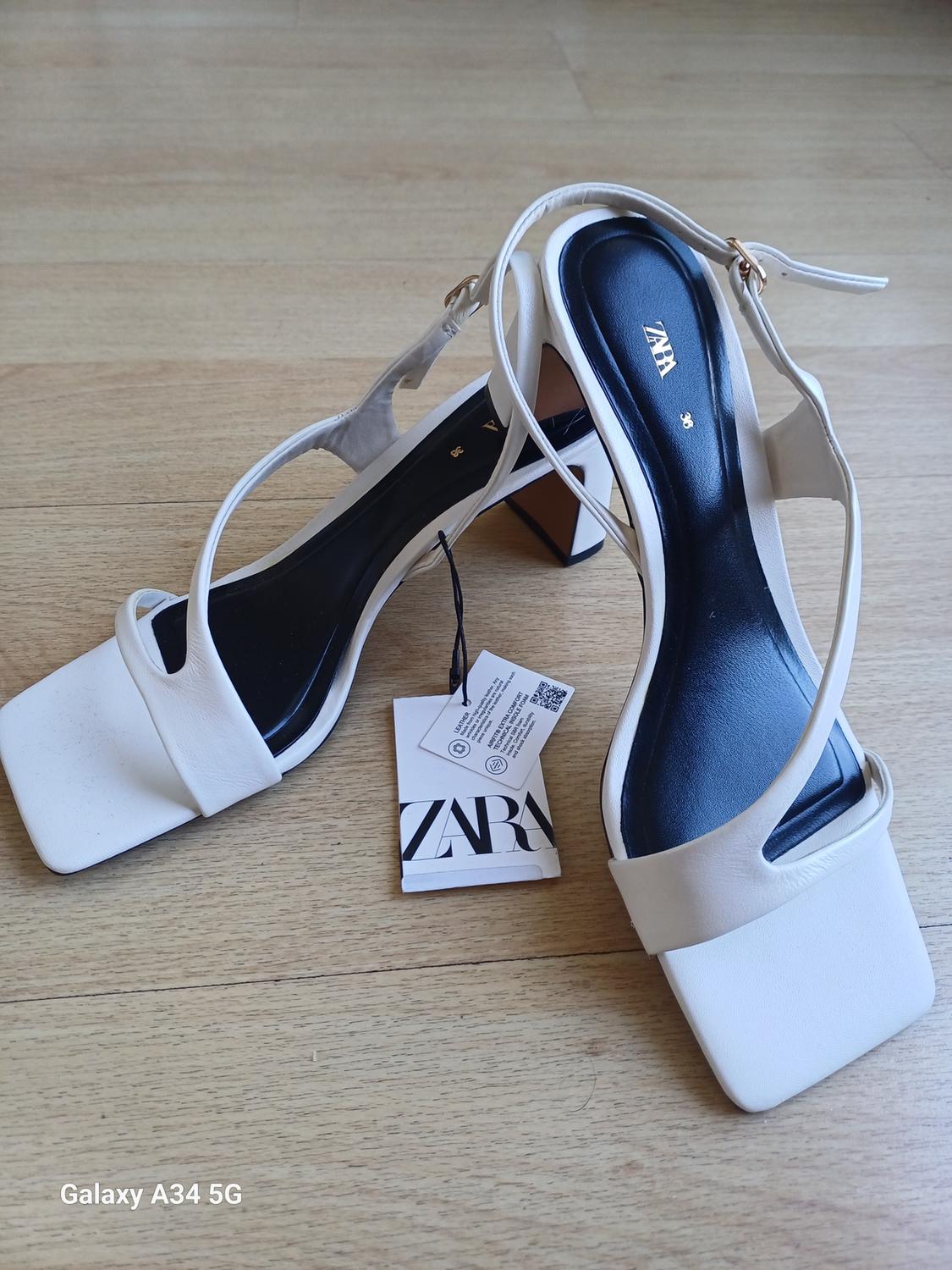 White block-heeled sandals with a black insole and Zara tag on a wooden floor. Visible text: "Galaxy A34 5G".