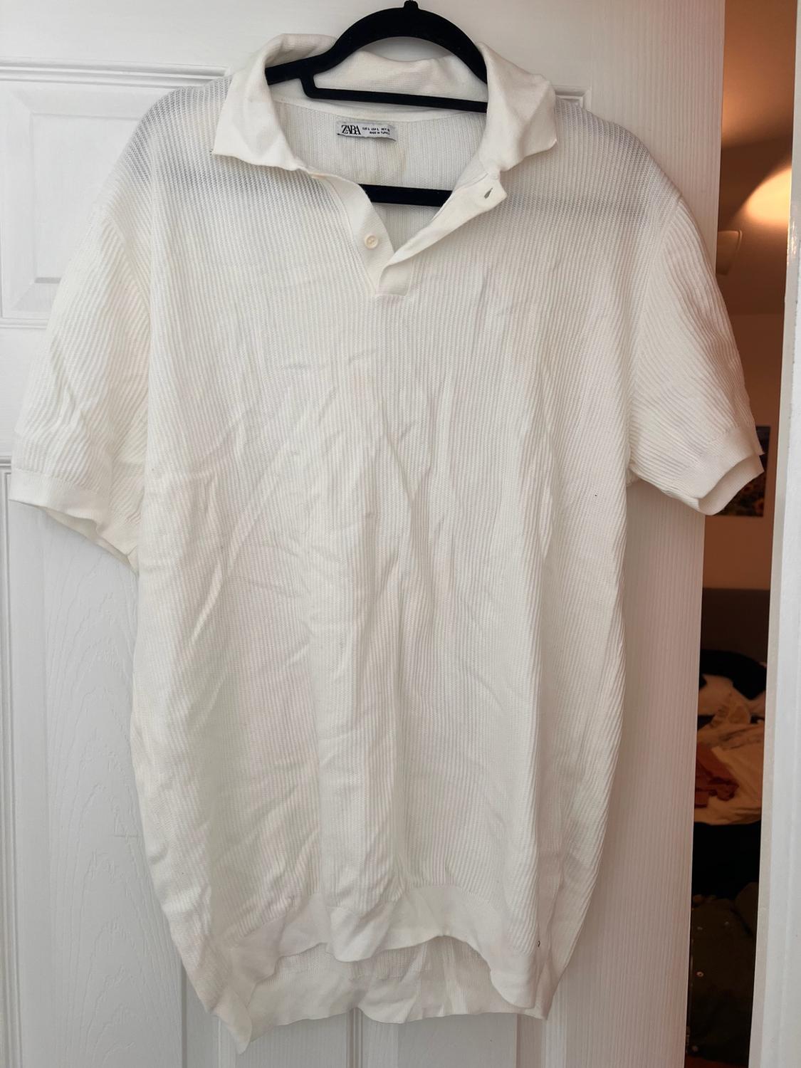 White short-sleeved textured polo shirt hanging on a black hanger on a white door. Label with text ZARA. Relaxed home setting.
