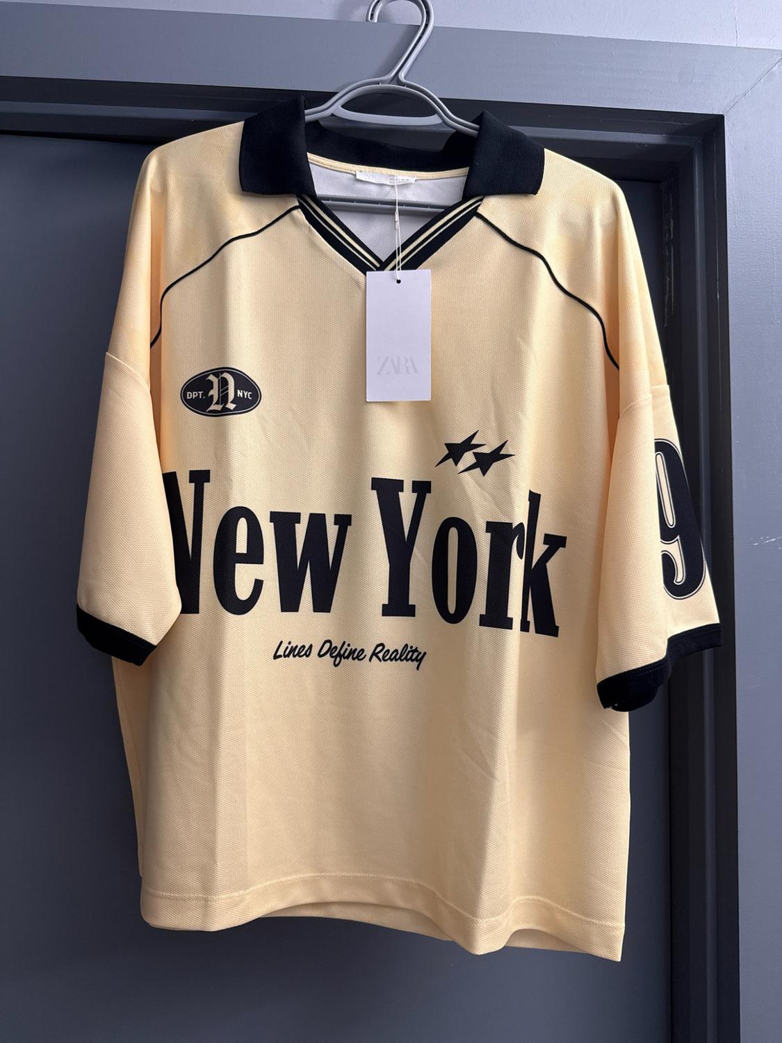 Yellow short-sleeved t-shirt on a hanger. Text: ZARA, DPT. N NYC, New York, Lines Define Reality, 9. Black collar, stars and grey background.