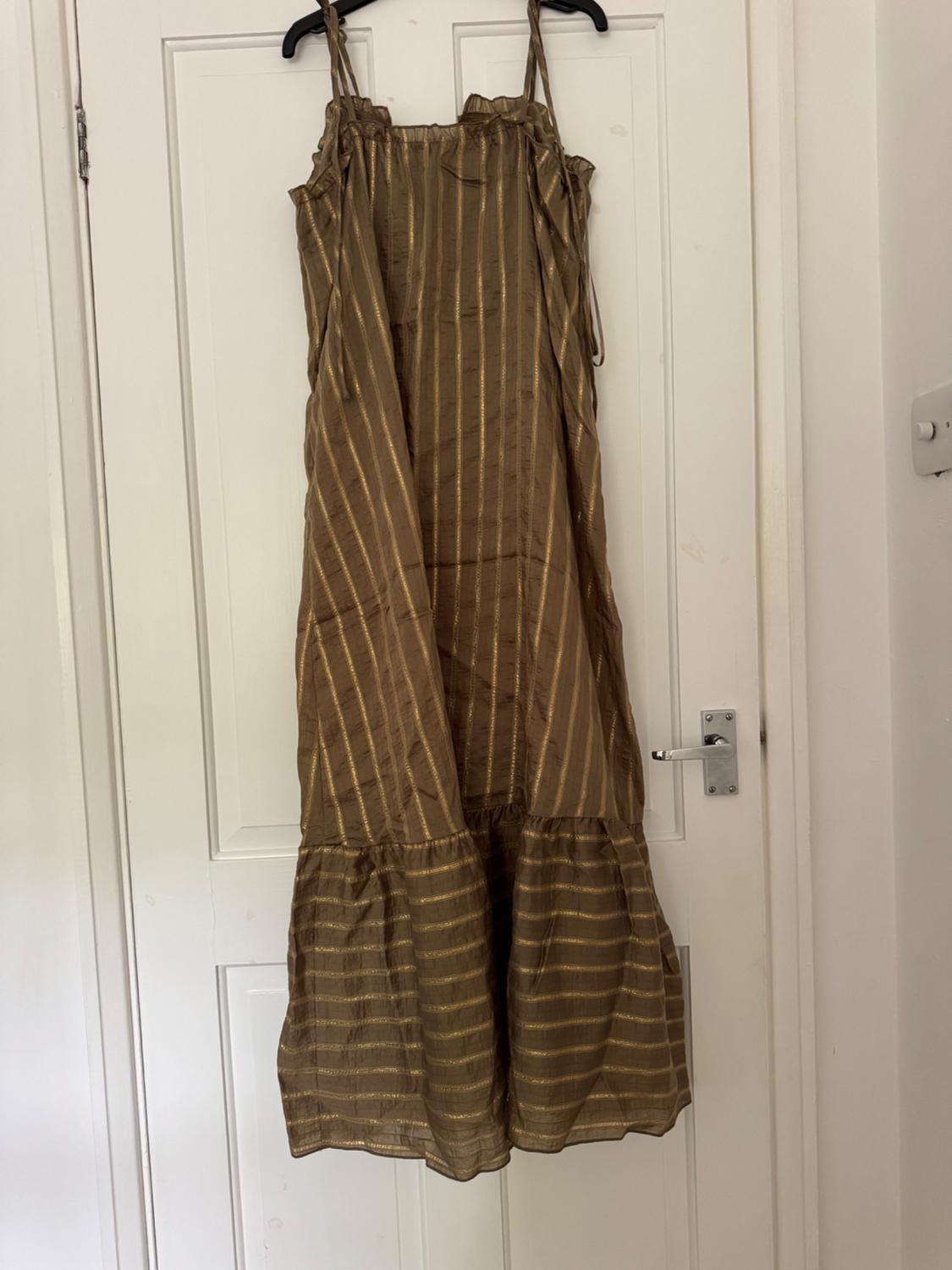 Brown strappy maxi dress with gold stripes, hanging on a white door in a relaxed and casual interior setting.