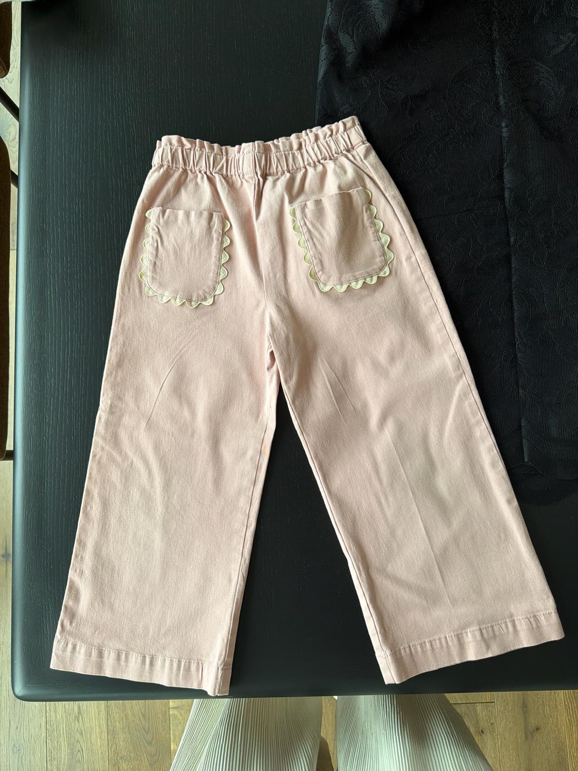 Pink pants with an elastic waistband and back pockets with contrasting scalloped trim, on a black table.