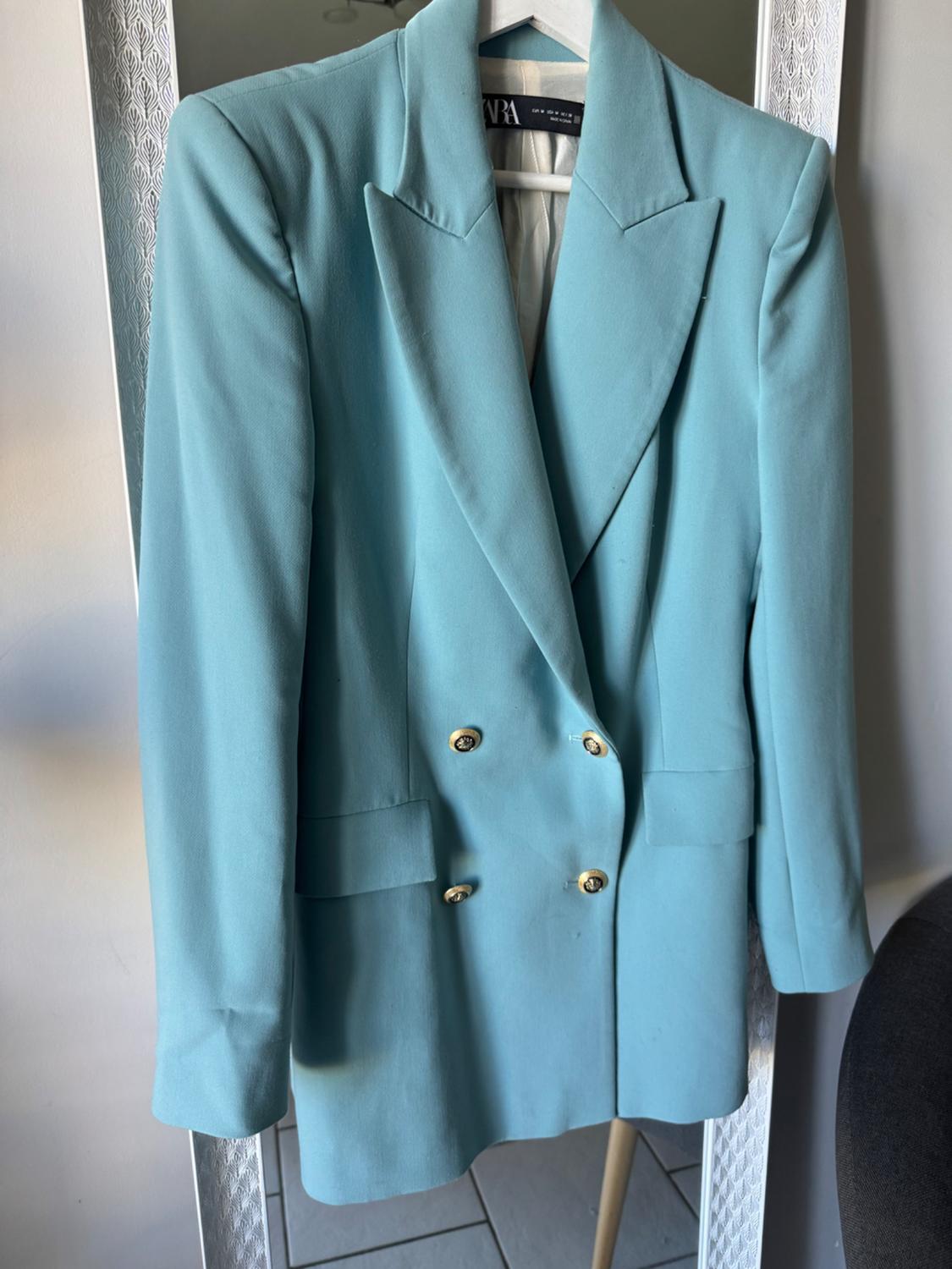 Light blue double-breasted jacket with gold buttons, hanging on a hanger in front of a decorative framed mirror.