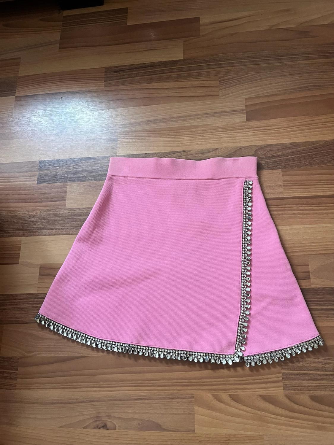 Pink mini skirt with sparkly rhinestone trim on the hem and front slit, placed on a wooden floor.
