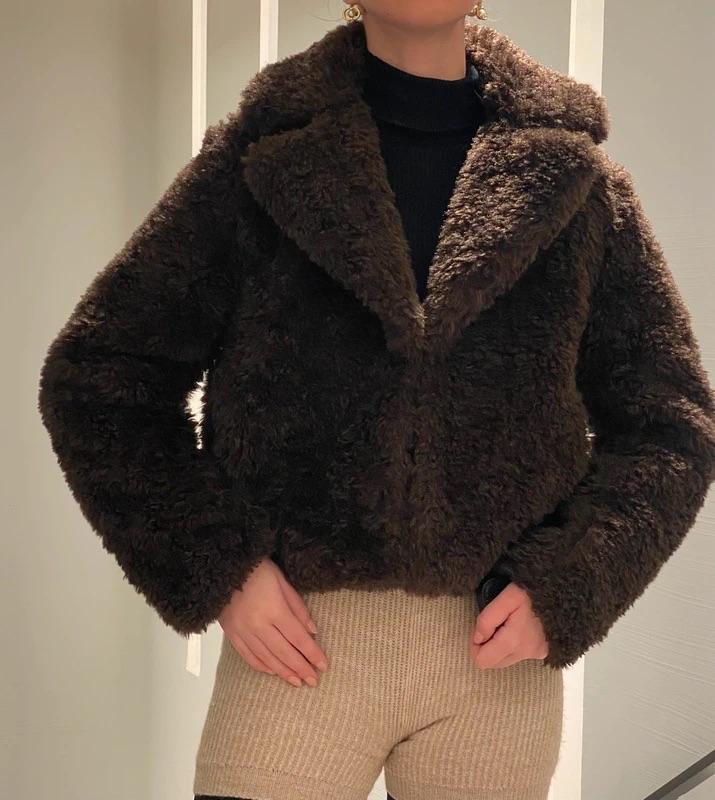 Model wearing a brown teddy jacket, black jumper, and beige shorts. Neutral background with light vertical lines.