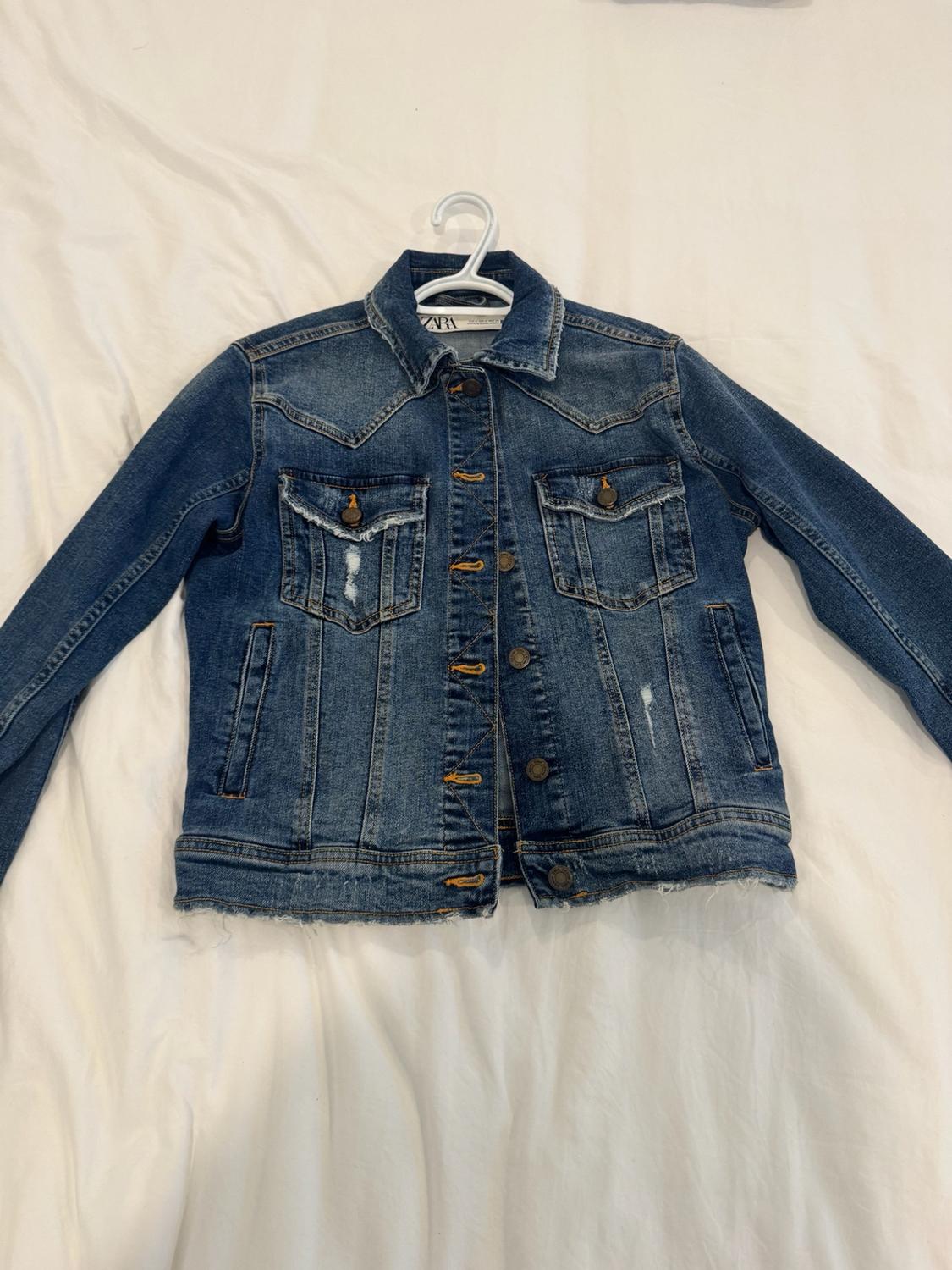 Blue denim jacket with pockets and distressed details on a hanger over a white sheet. Label with ZARA text.