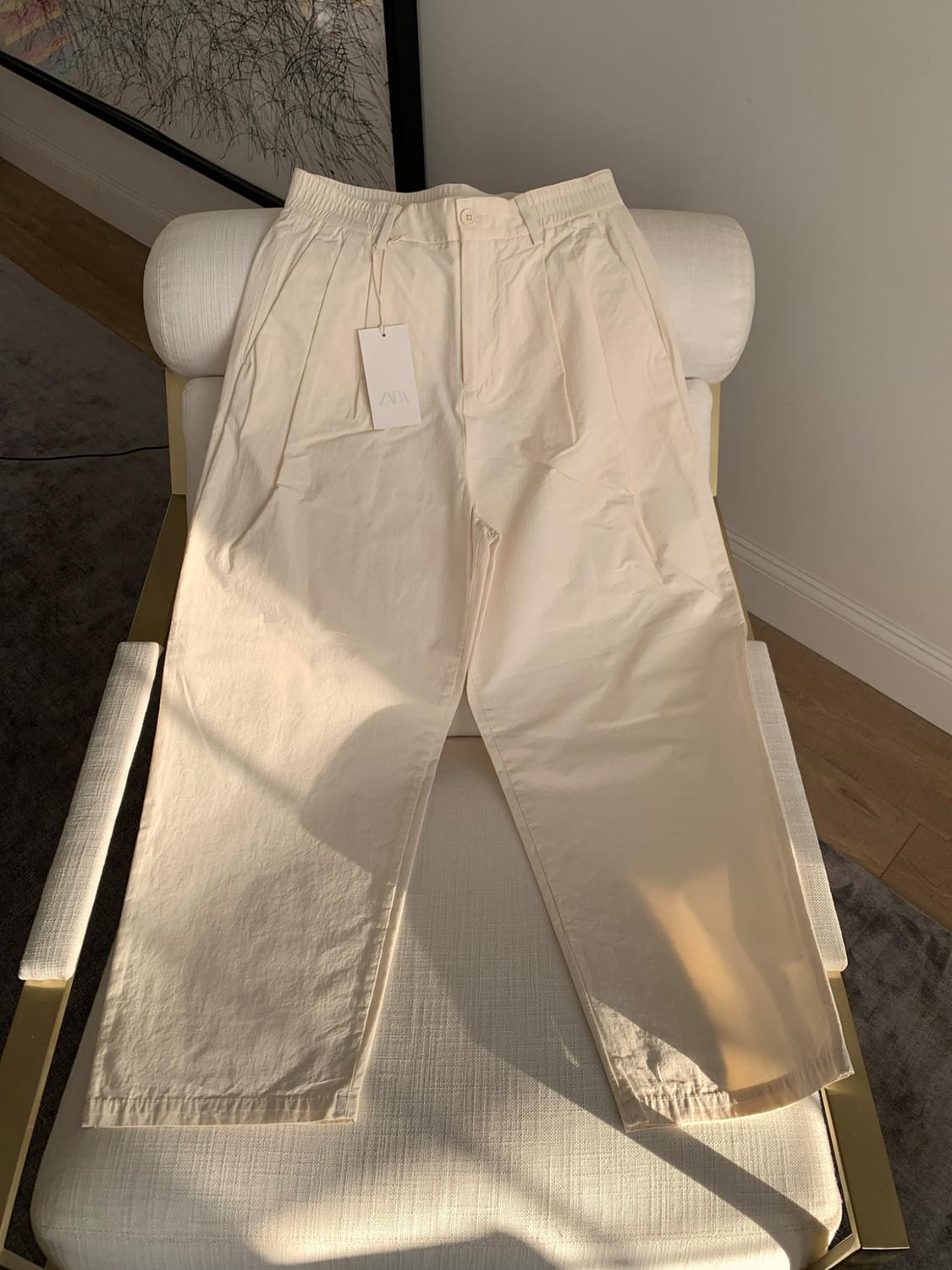 Beige pleated trousers with a ZARA label on a white chair. Natural light and sharp shadows create a relaxed atmosphere.