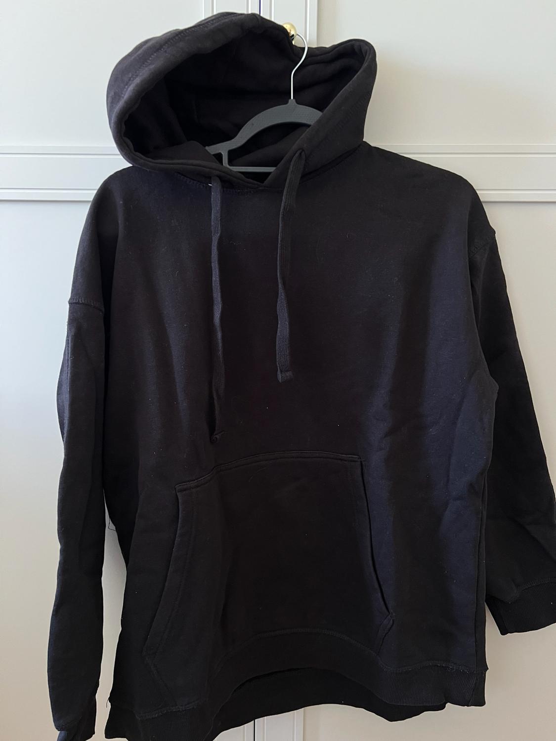 Black hoodie with drawstring and kangaroo pocket, hanging on a hanger on a white door. Casual and relaxed style.