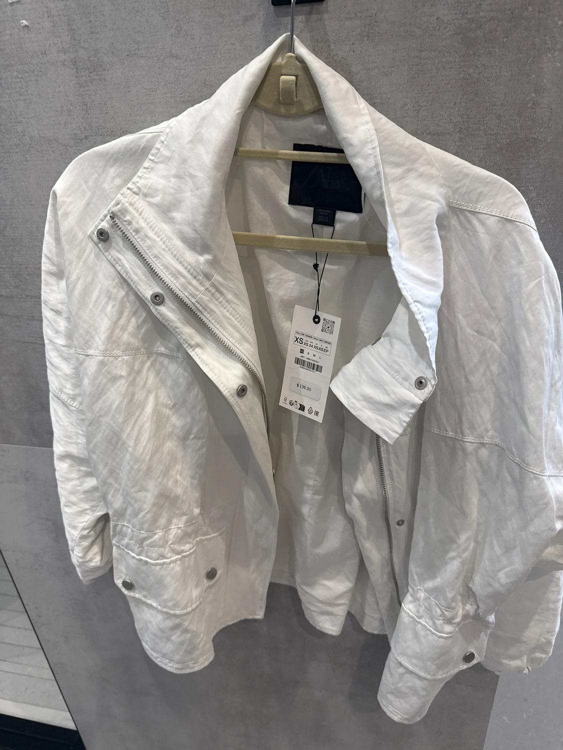 White jacket with a zipper and pockets hanging on a hanger against a gray background. Text: XS, S M L, $139.00.