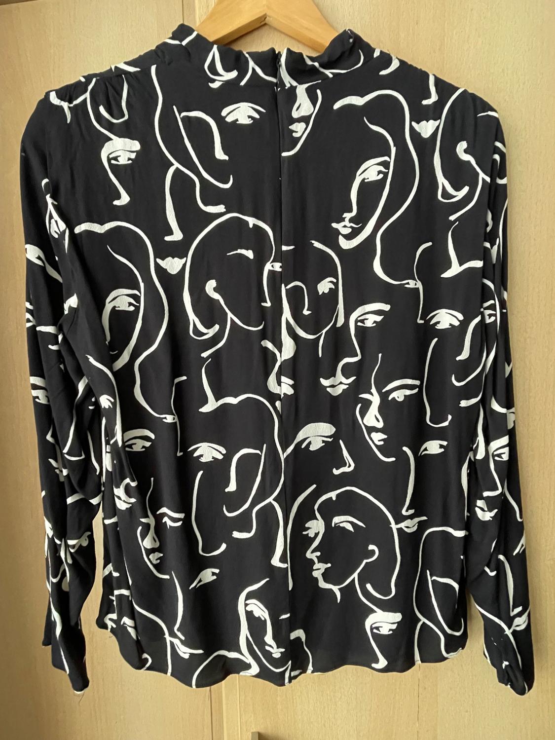 Black shirt with white abstract face print, hanging on a wooden hanger against a light wood background.
