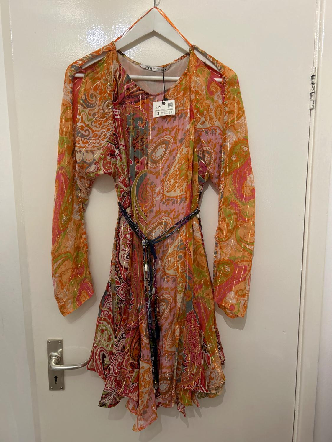 Multicoloured patterned dress with long sleeves, hanging on a white hanger over a door. Visible label with size S.