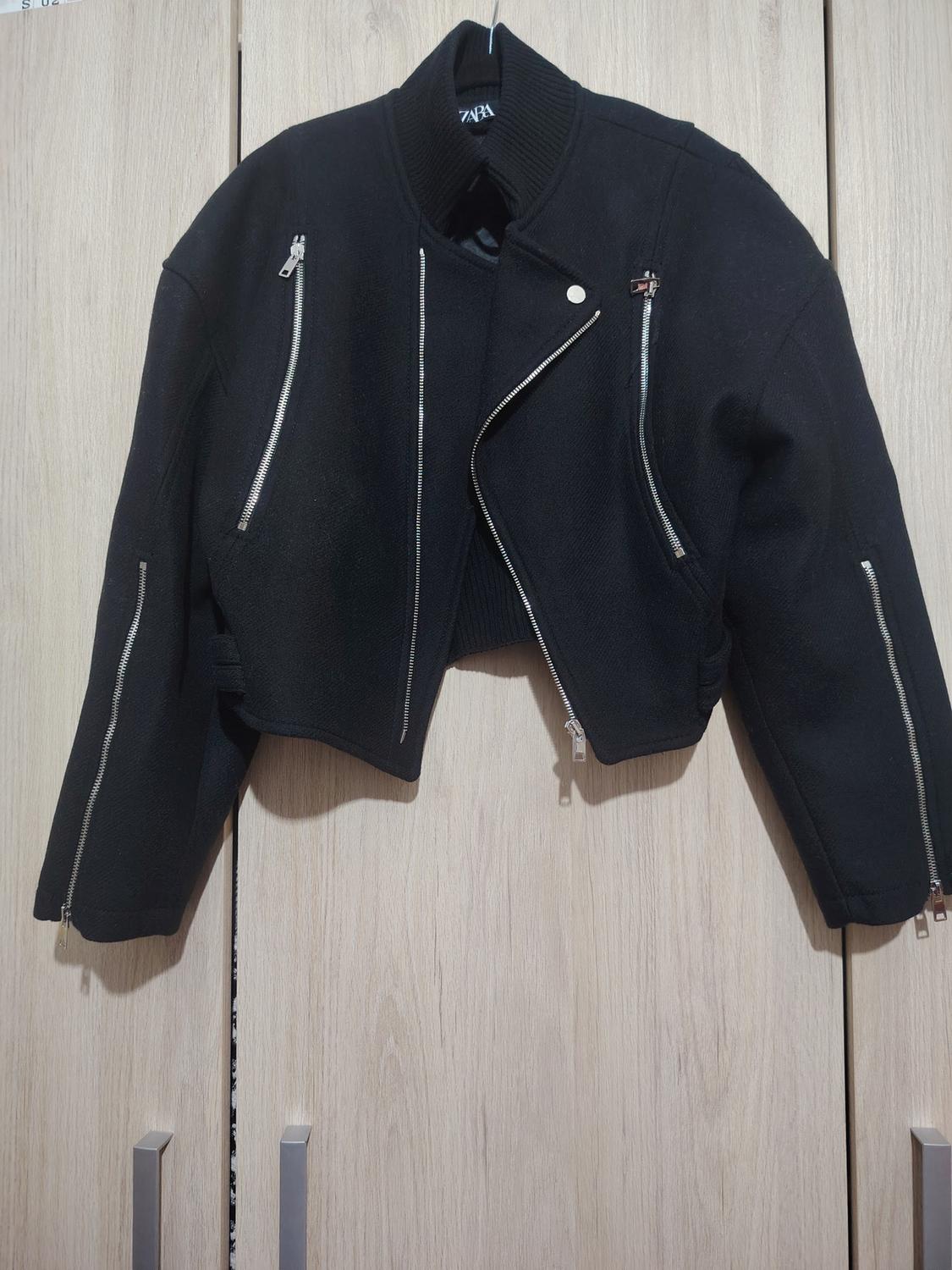 Black jacket with silver zips hanging on a light wooden door. Visible label on the collar.