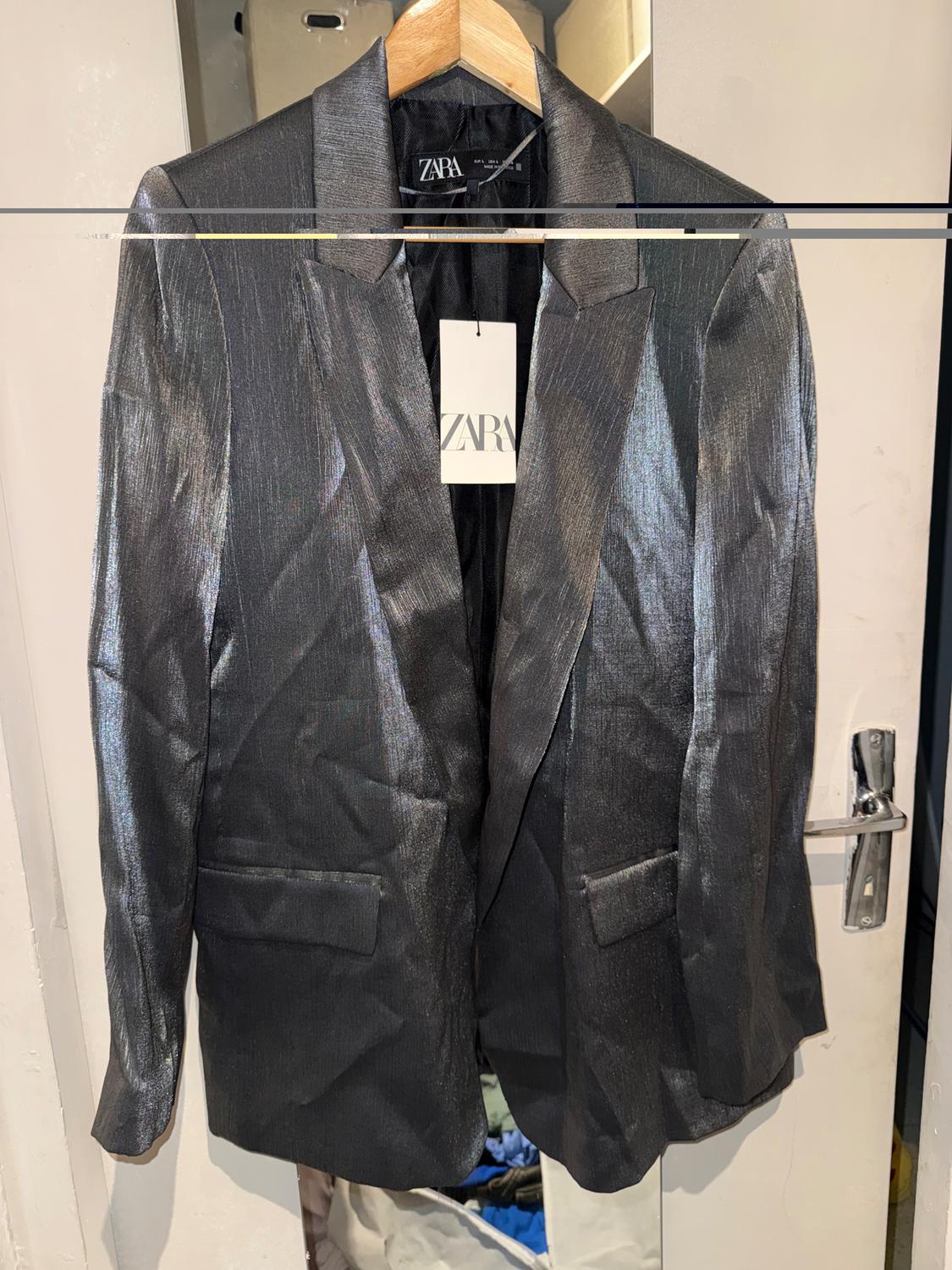 Grey metallic jacket hanging on a wooden hanger. Visible label with the text "ZARA". Background of a door and white wall.