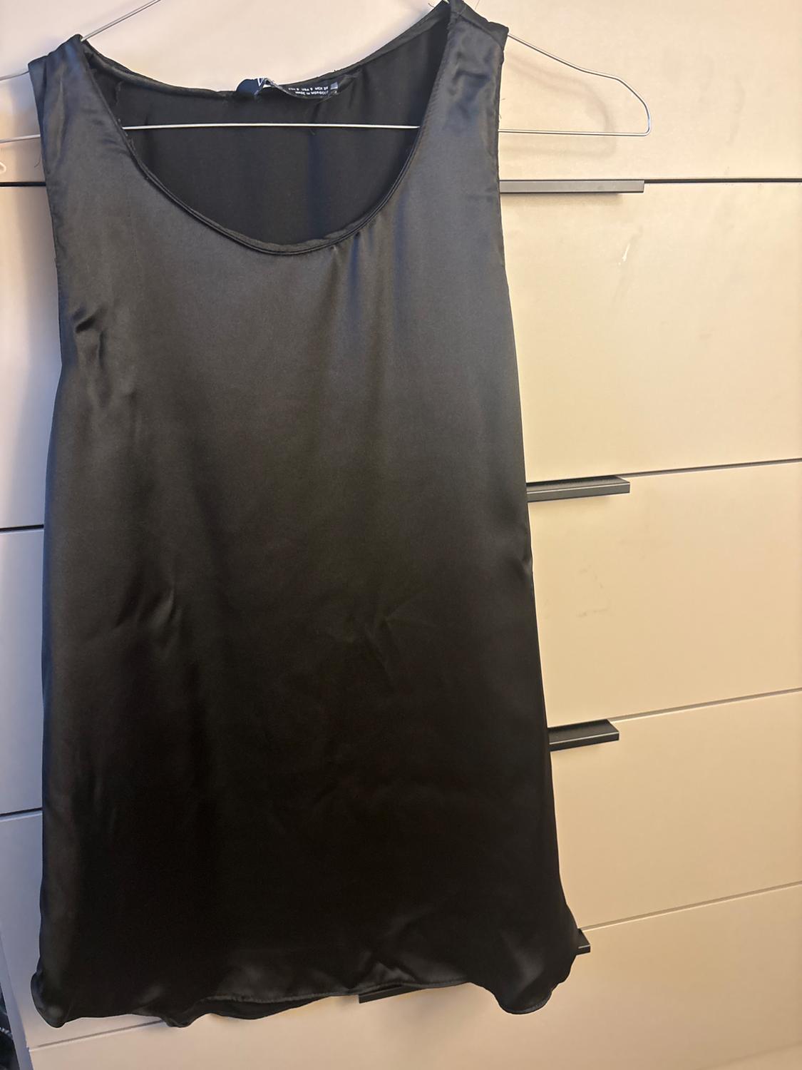 Sleeveless black satin dress hanging on a hanger in front of a background of light-coloured drawers.