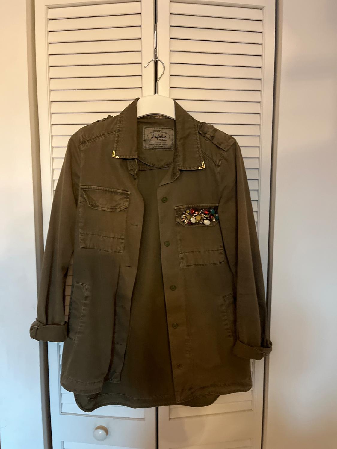 Olive green jacket hanging on a hanger, with pockets and diamante details on the upper left pocket, white door background.