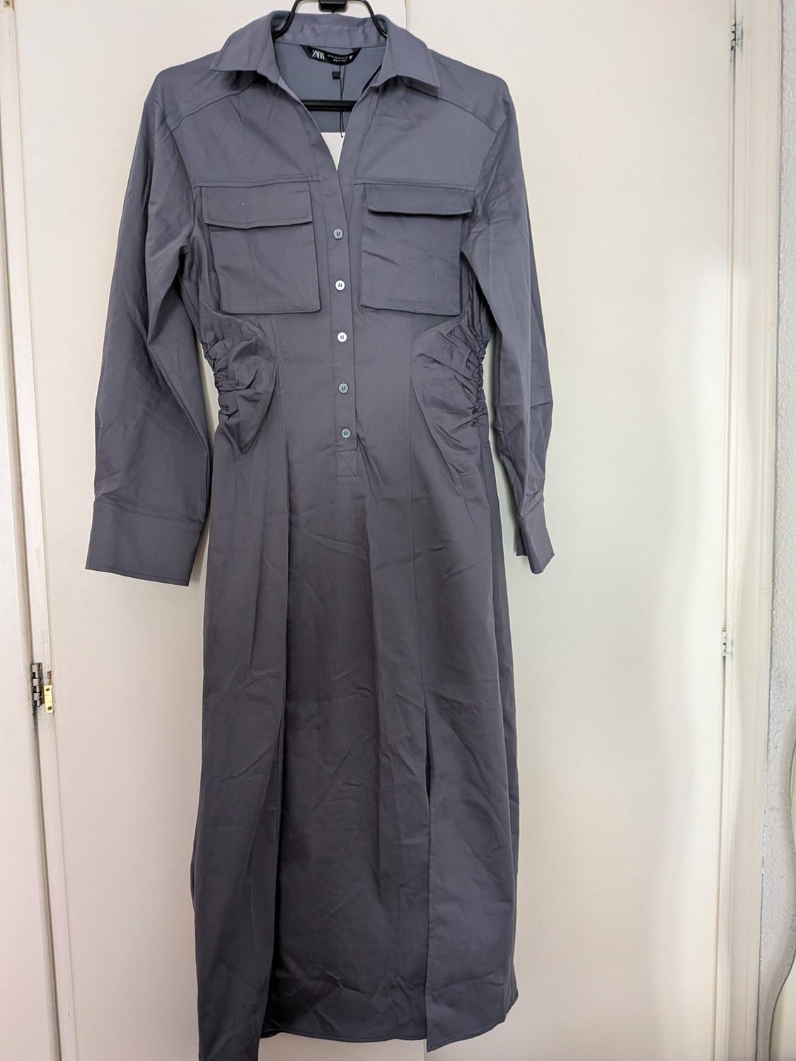 Blue-grey shirt dress hanging on a white door. ZARA label. Long sleeves, pockets, gathered waist and front opening.