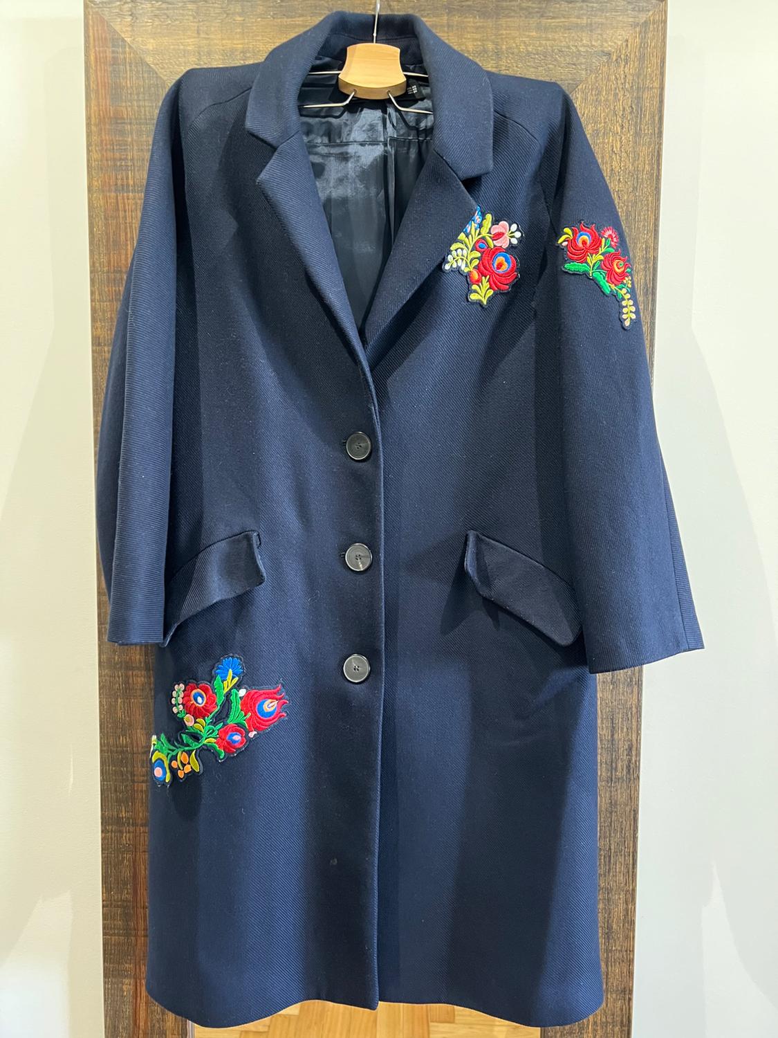 Dark blue coat with colourful floral embroidery on the chest, sleeve, and pocket, hanging on a wooden hanger.