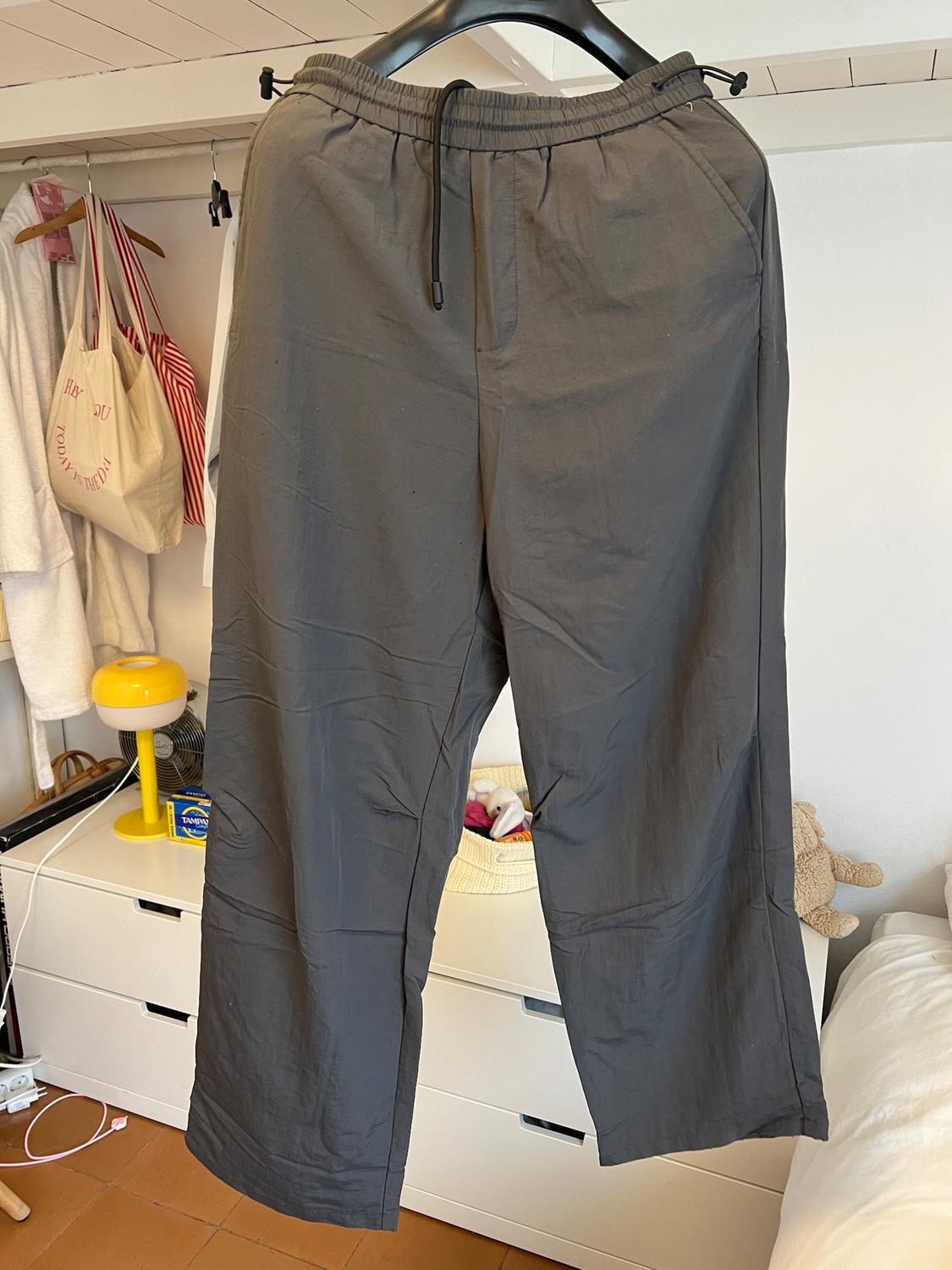 Grey trousers hanging on a hanger against a bedroom background with a white chest of drawers, yellow lamp, and hanging bags.