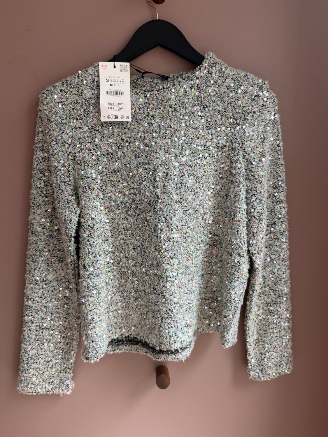 Grey textured-knit jumper with sequins hanging on a black hanger against a pink wall. Text: S US $49.90 UK £29.90.