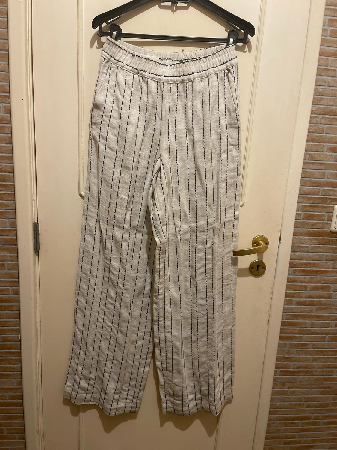 Beige wide-leg trousers with black vertical stripes and an elasticated waist, hanging on a black hanger over a white door.