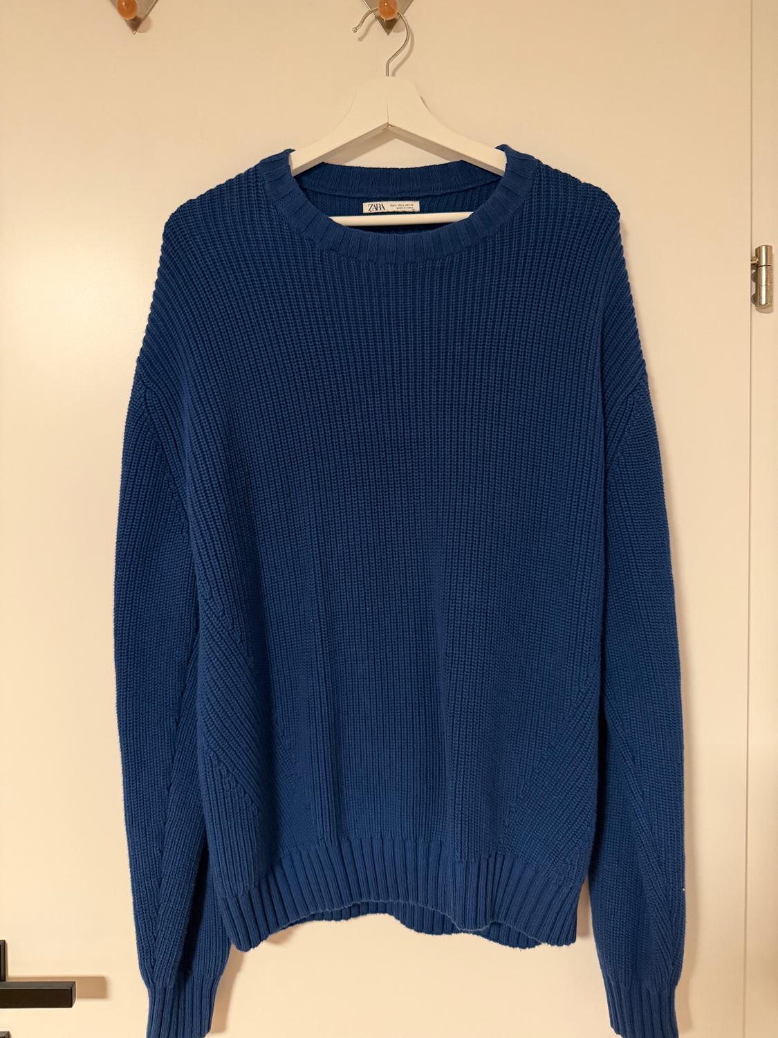 Blue chunky knit jumper hanging on a white hanger over a light door. Label visible on the collar.